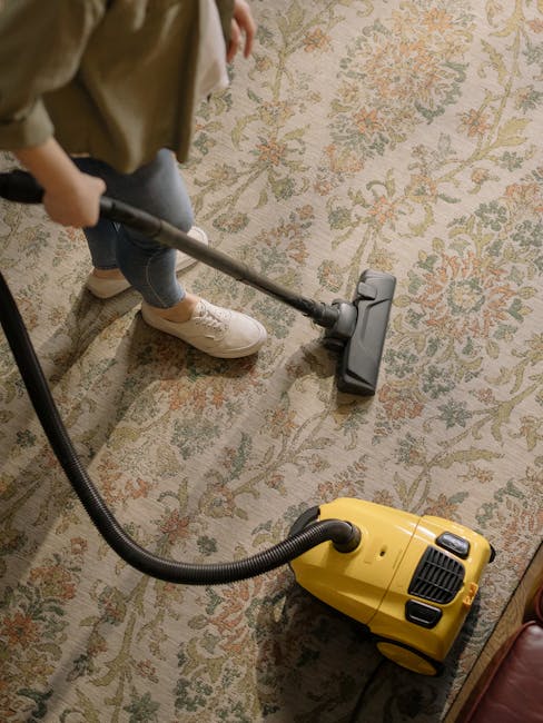 Close-up view of a beige and cream-colored carpet with a textured pattern, showing areas of dust and dirt accumulation, illuminated by natural light coming from the side. The image highlights the impo
